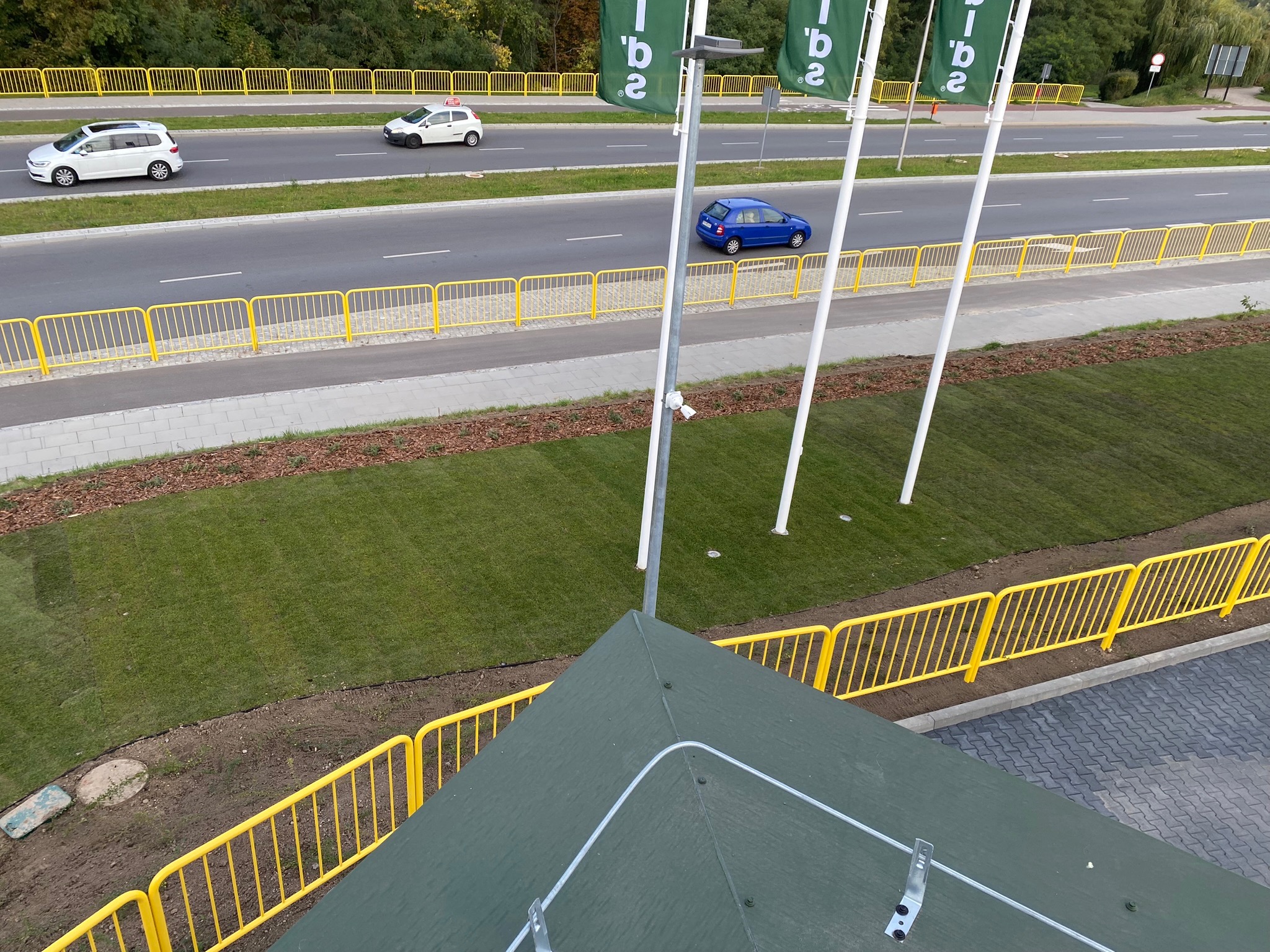 Widok z góry na świeżo założony trawnik z rolki, oddzielony żółtymi barierkami od drogi z samochodami, z widocznym dachem budynku w dolnej części kadru.