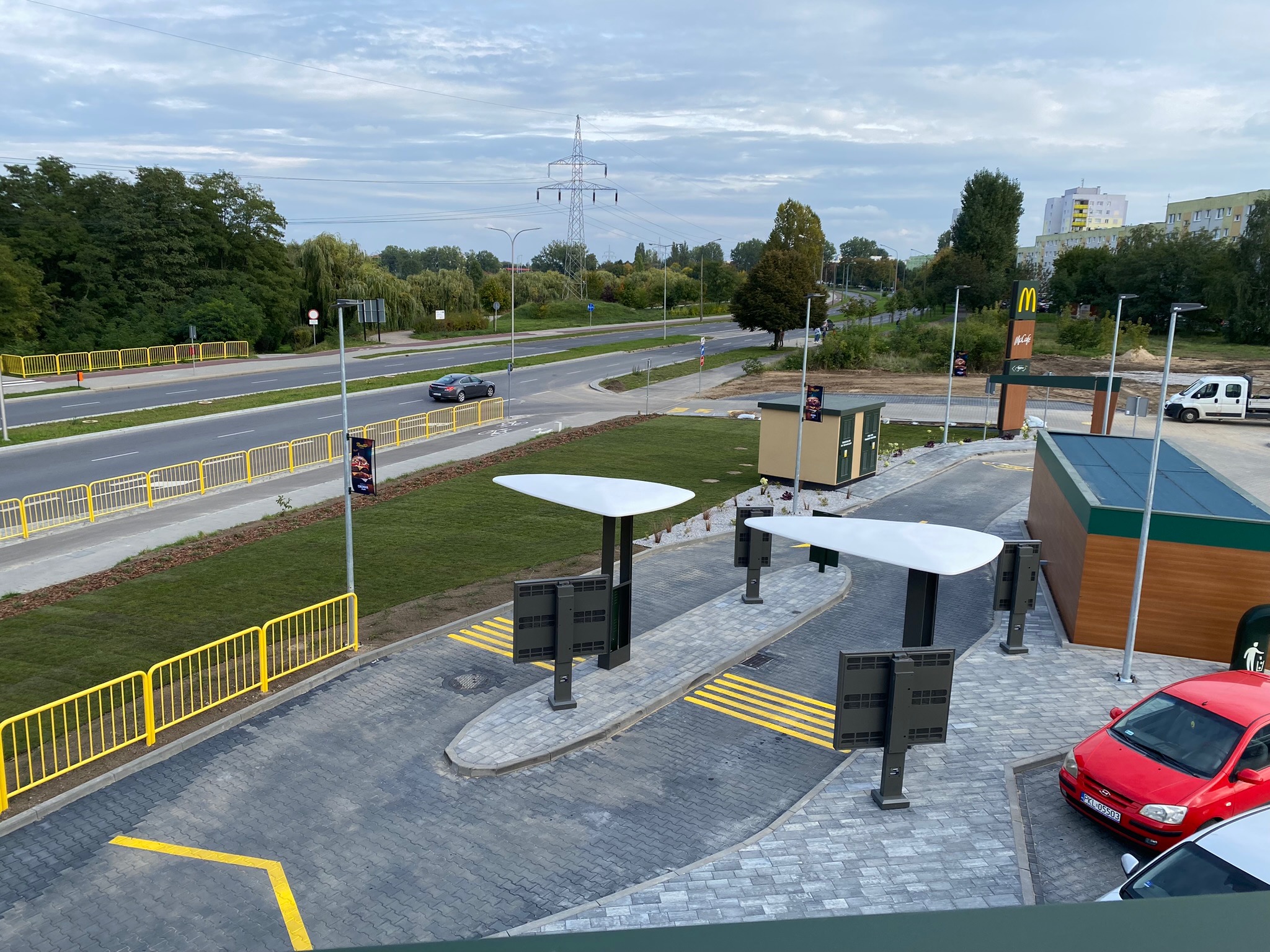 Widok z góry na nowo zagospodarowany teren wokół restauracji typu drive-thru, z widocznym świeżo posianym trawnikiem, elementami małej architektury i fragmentem drogi z żółtymi barierkami.