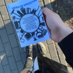 Ulotka reklamowa z napisem 'Mobilne Sprzątanie' i ilustracjami narzędzi czyszczących, trzymana w dłoni na tle chodnika i fragmentu stylizowanej torebki.