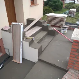 Przygotowanie schodów wejściowych do domu pod montaż płytek: widoczne ułożone płytki na zaprawie, poziomica, paczka płytek oparta o ścianę, wiadro z zaprawą i różowe buty leżące na górnym stopniu.