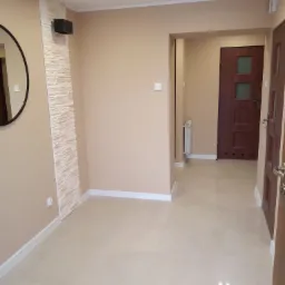 Korytarz po remoncie z beżowymi ścianami, jasnymi płytkami podłogowymi, dekoracyjną ścianą z kamienia i okrągłym lustrem.