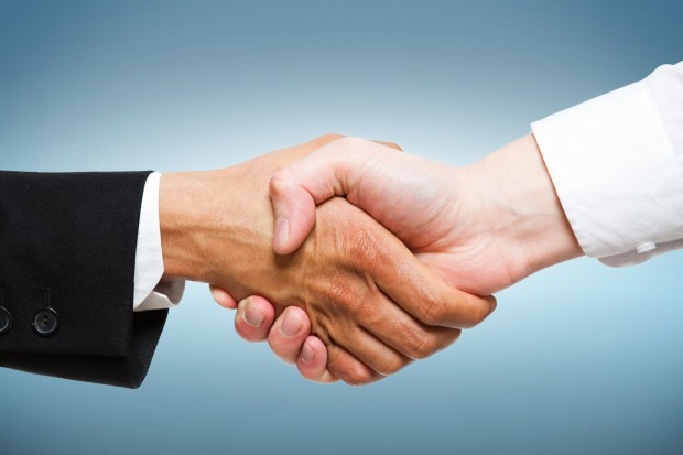 Uścisk dłoni: jasna dłoń w białej koszuli ściska ciemniejszą dłoń w czarnym garniturze na tle gradientu błękitu.