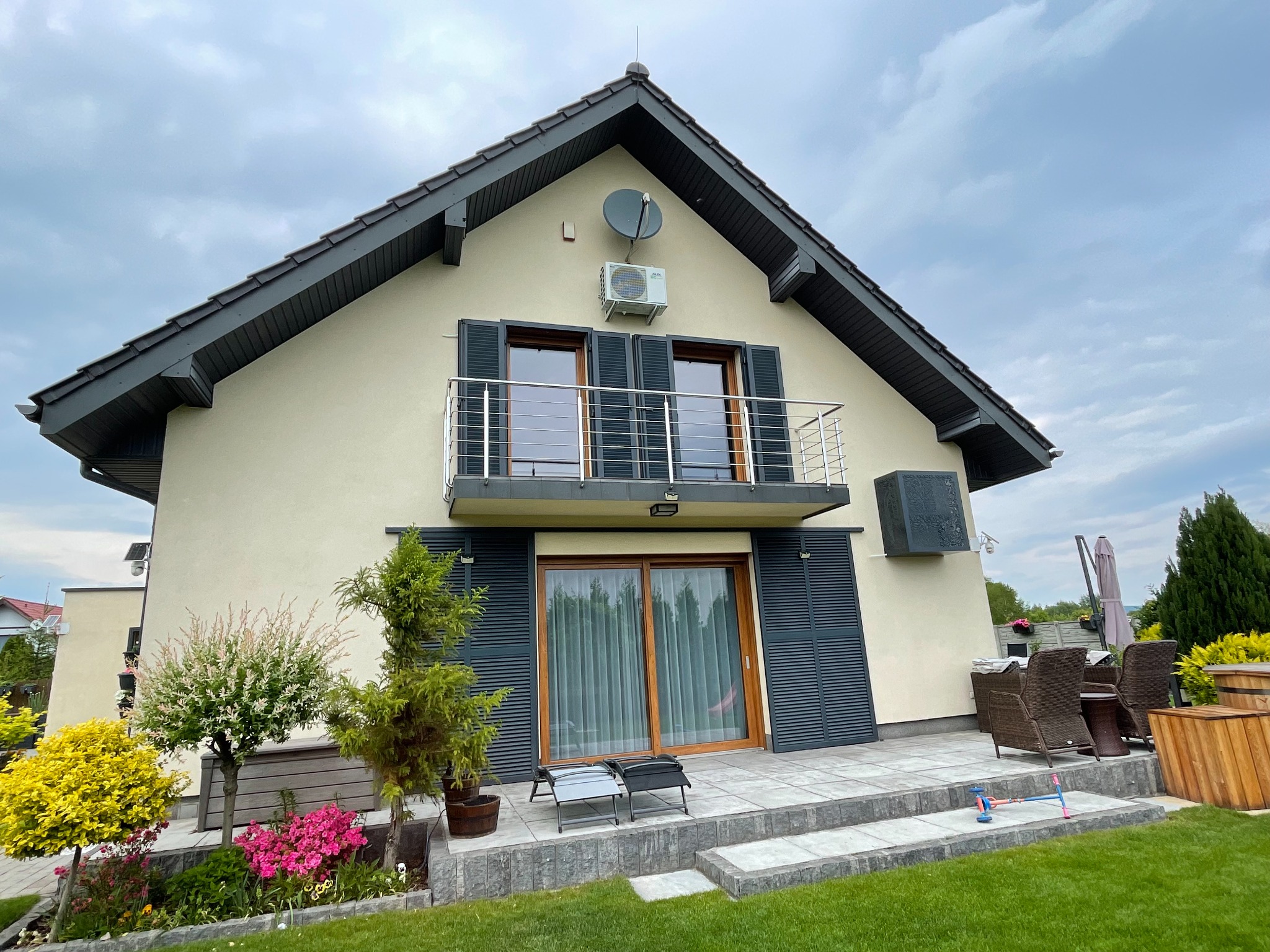 Jasny dom jednorodzinny z zamontowaną jednostką zewnętrzną klimatyzacji AUX na fasadzie, widoczna antena satelitarna powyżej, okna z ciemnymi okiennicami, balkon z metalową balustradą, ogród...