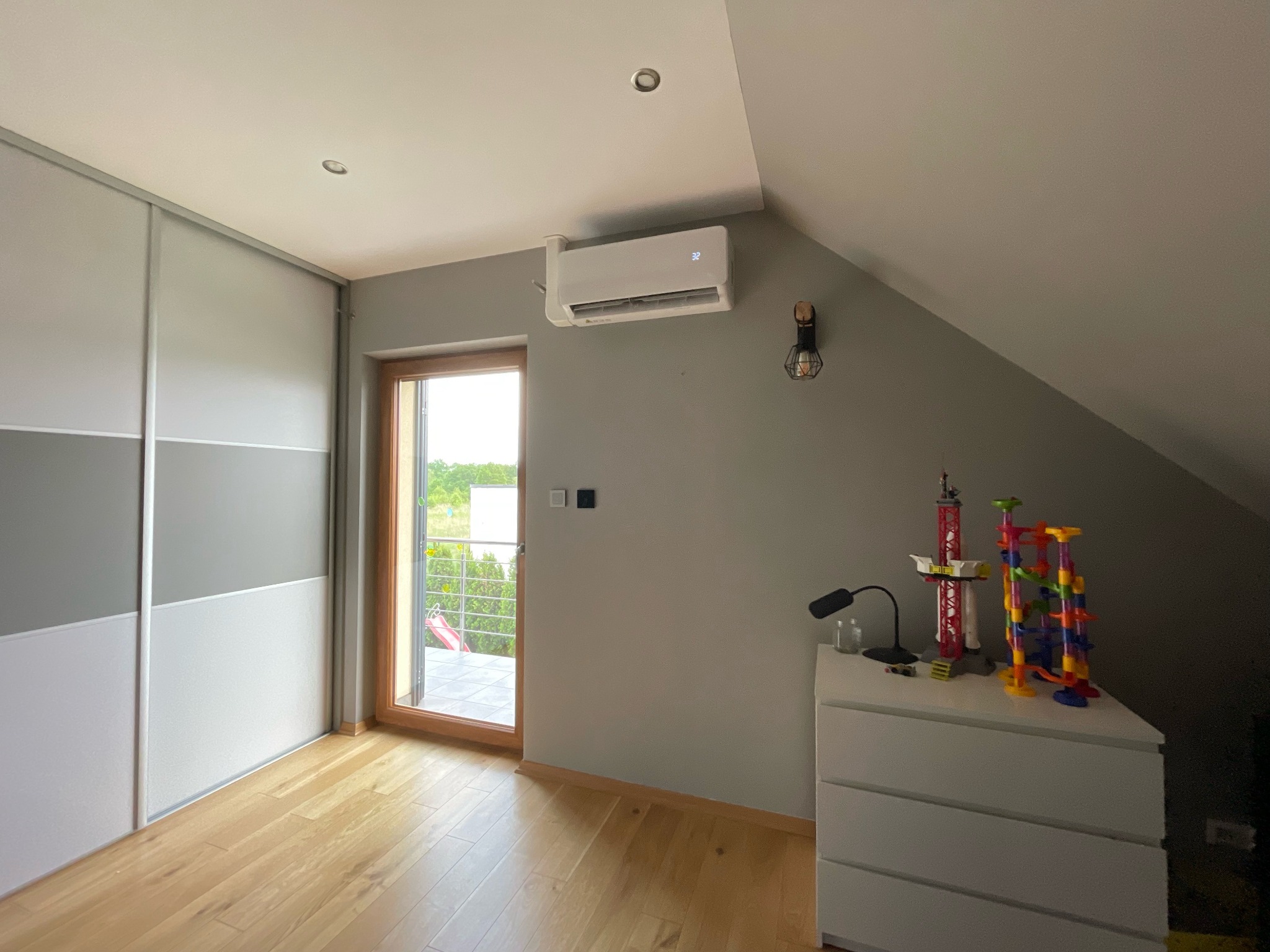Biała klimatyzacja ścienna zamontowana w pokoju na poddaszu z drewnianą podłogą, obok okna balkonowego i szafy przesuwnej, na komodzie lampka i zabawki dziecka.