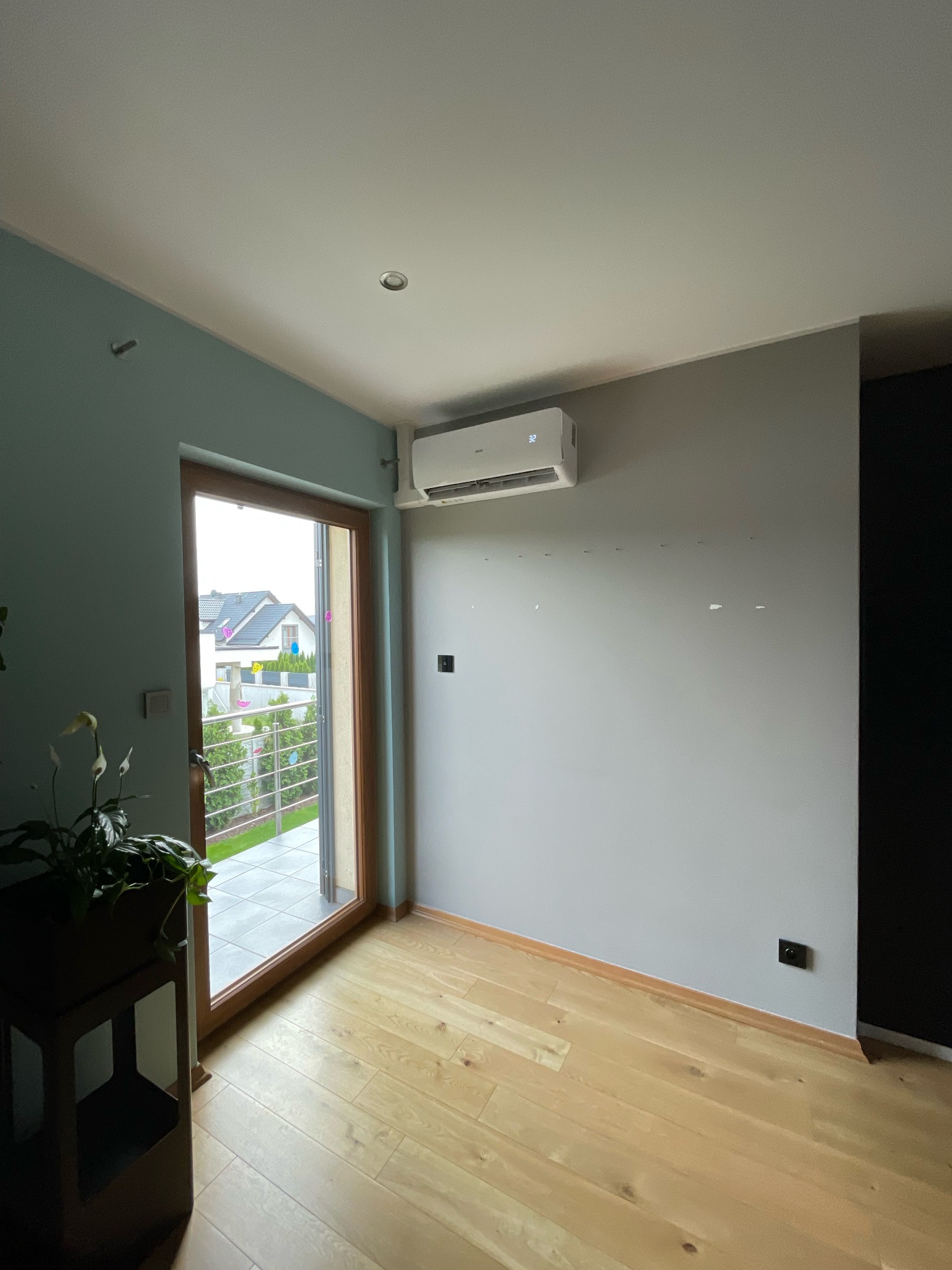 Wnętrze pokoju z zamontowaną klimatyzacją ścienną, widoczne ślady po poprzednich mocowaniach na ścianie, parkiet na podłodze i wyjście na balkon.