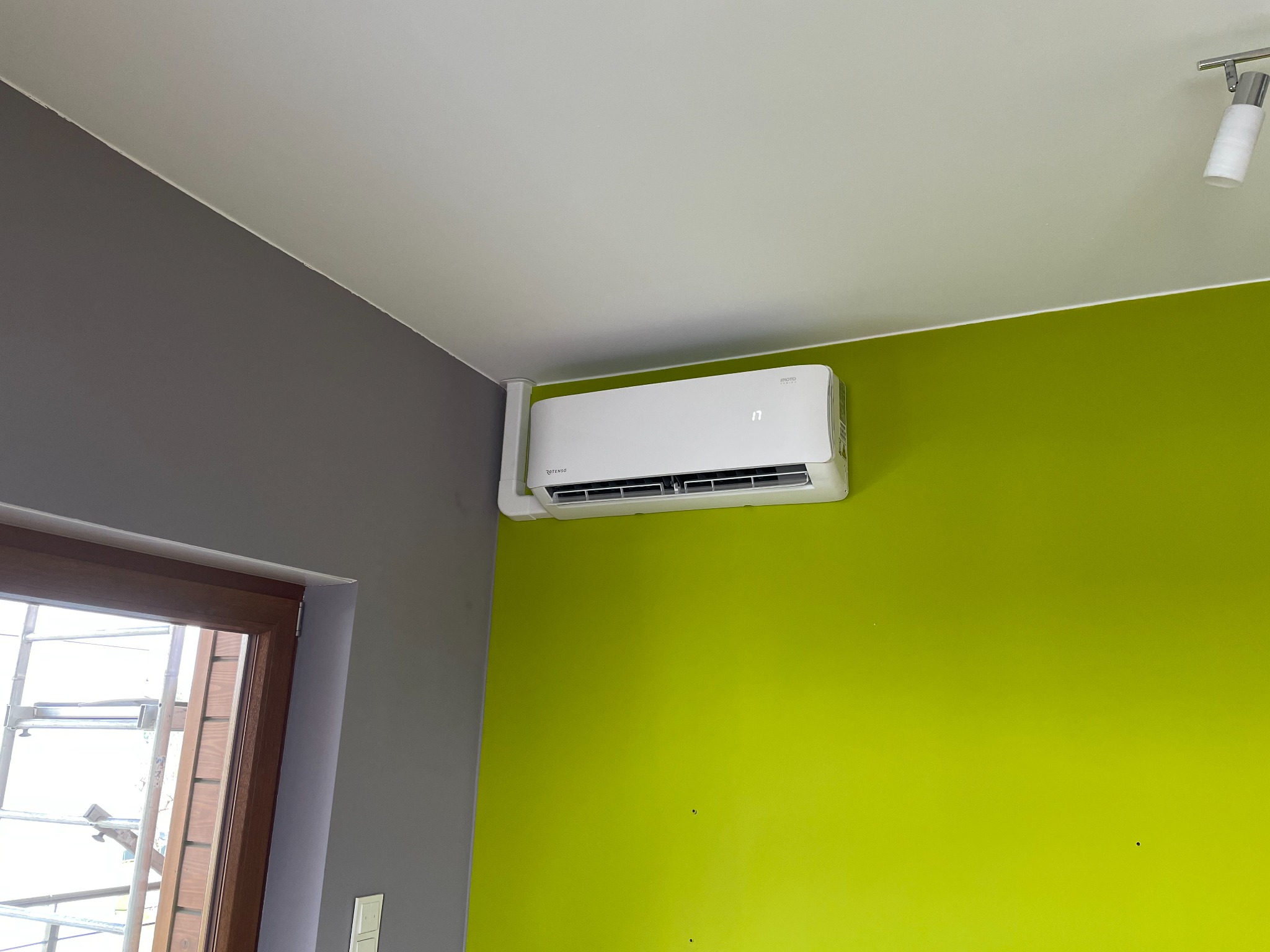 Biały klimatyzator ścienny zamontowany w rogu pokoju z zieloną i szarą ścianą, widoczna lampa sufitowa i okno z rusztowaniem na zewnątrz.