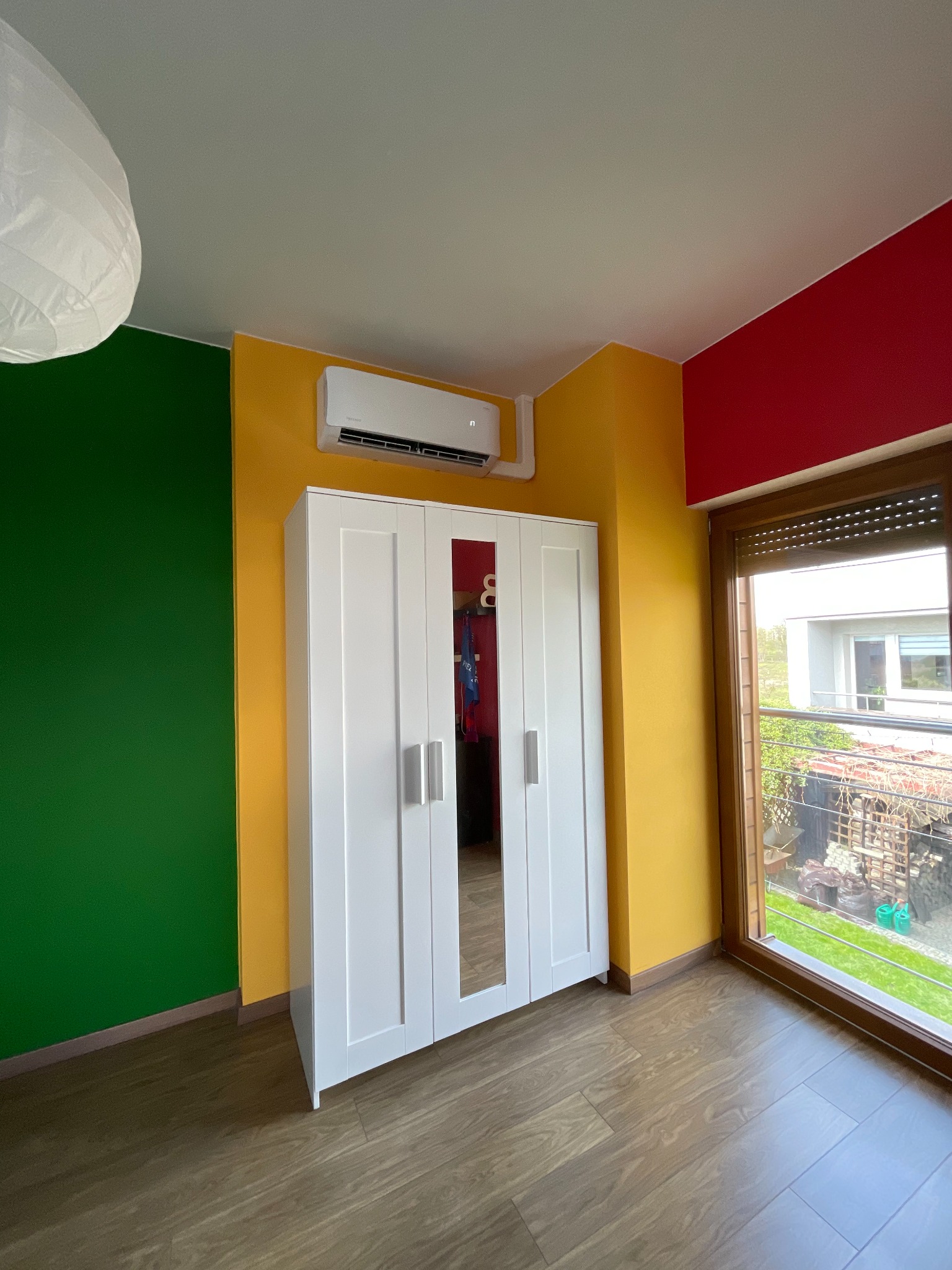 Biały klimatyzator ścienny zamontowany nad białą szafą w pokoju z zieloną, żółtą i czerwoną ścianą, widok z okna na zewnątrz.
