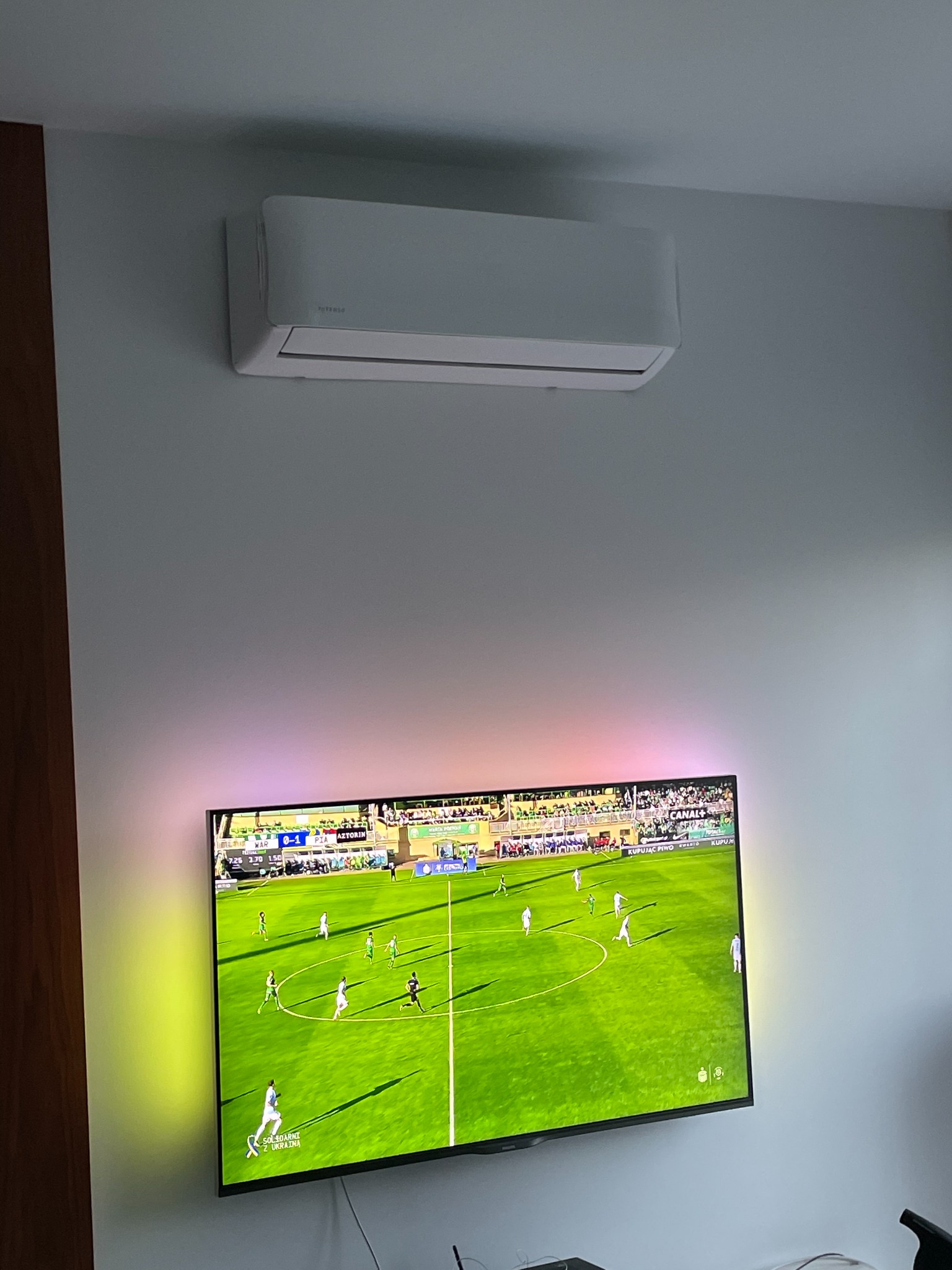 Biały klimatyzator ścienny zamontowany nad telewizorem z podświetleniem LED, na którym wyświetlany jest mecz piłki nożnej.