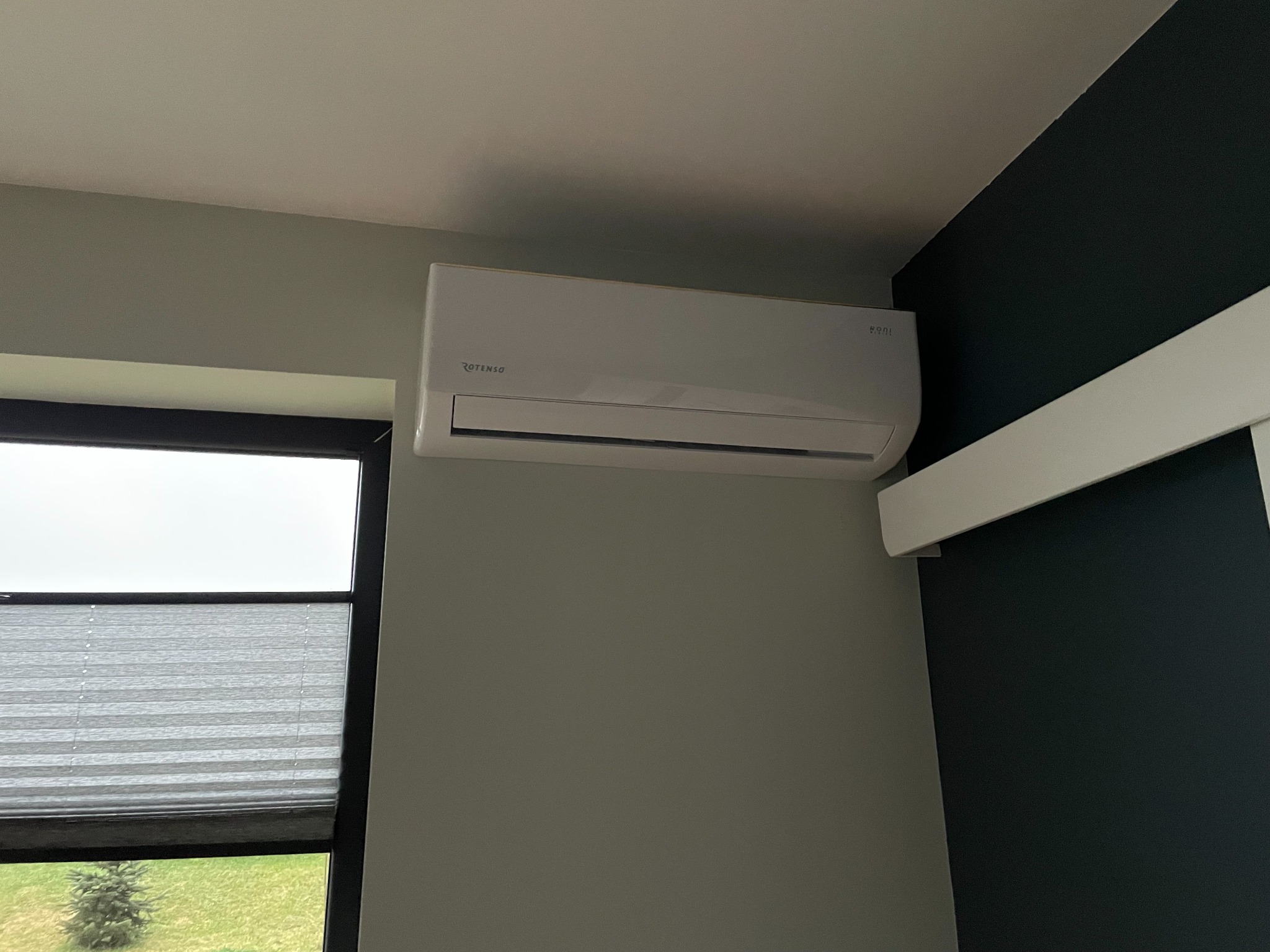 Biały klimatyzator Rotenso zamontowany na ścianie w pokoju z oknem i roletami plisowanymi.