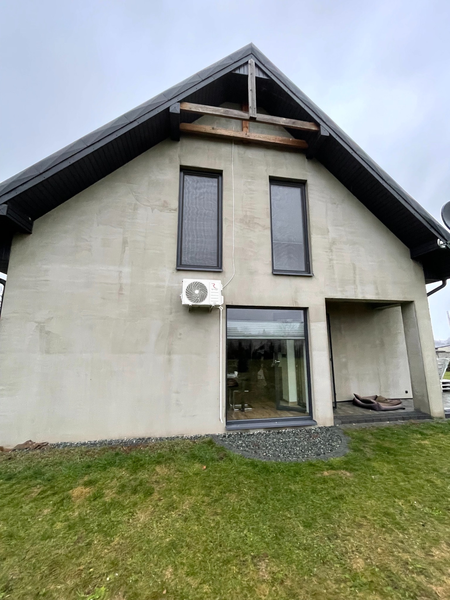 Szary dom jednorodzinny z zamontowaną jednostką zewnętrzną klimatyzacji na ścianie, widok z trawnika.