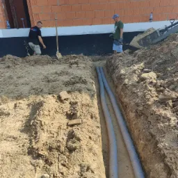 Dwaj mężczyźni pracują przy wykopie pod budynkiem z cegły, w którym ułożone są dwie rury w osłonie, widoczna łopata i sprzęt budowlany.