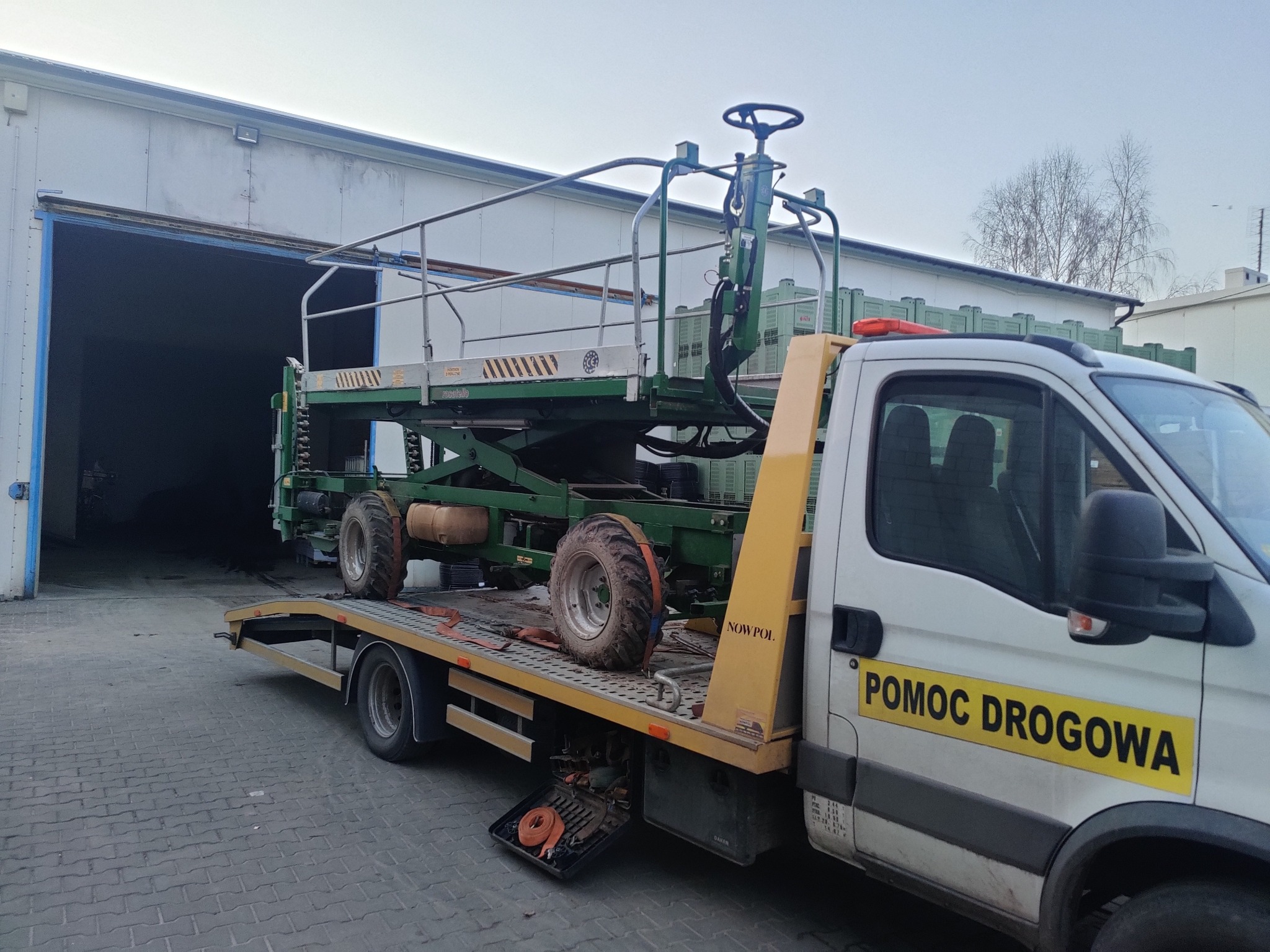 Laweta pomocy drogowej z podnośnikiem koszowym na platformie, w tle hala magazynowa z otwartą bramą.
