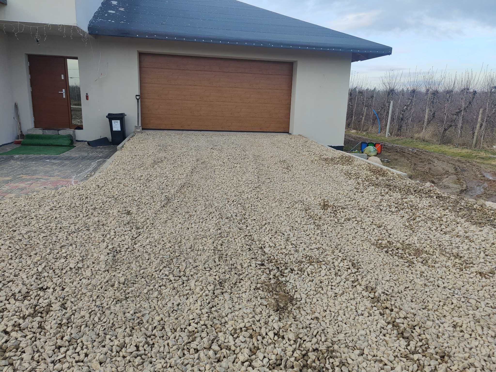 Podjazd do garażu wysypany jasnym żwirem, widoczny fragment domu z brązową bramą garażową i drzwiami wejściowymi, w tle sad drzew owocowych.