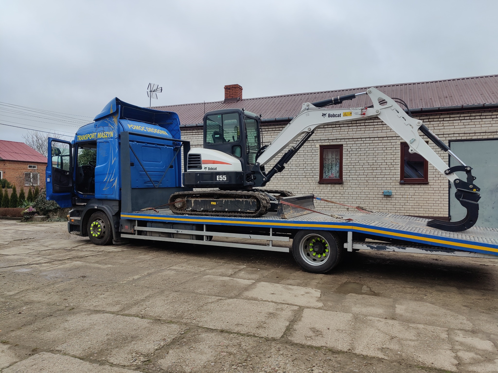 Niebieska laweta z minikoparką Bobcat E55 na platformie, zaparkowana na betonowym placu przed budynkiem z cegły.