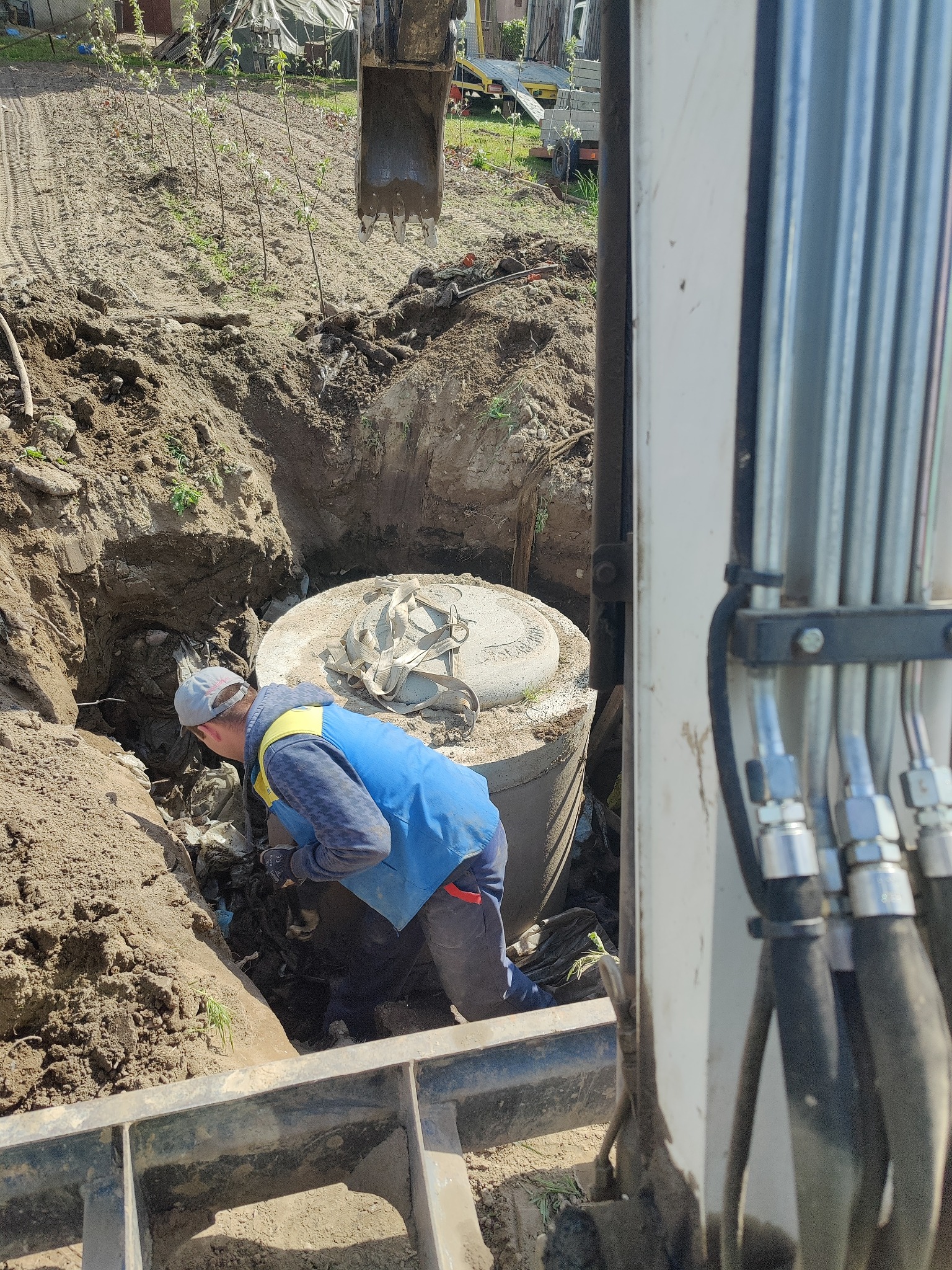 Pracownik w wykopie obok betonowego zbiornika z pasami mocującymi, widok z kabiny koparki, ziemia wokół wykopu.