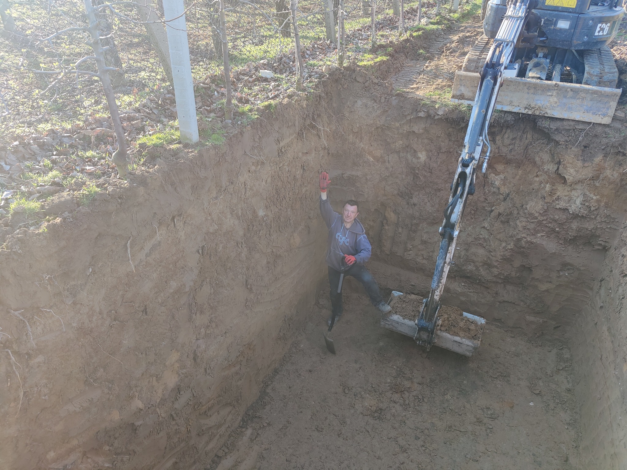 Głęboki wykop w ziemi z pracującą koparką i stojącym w dole mężczyzną z łopatą, w tle drzewa.