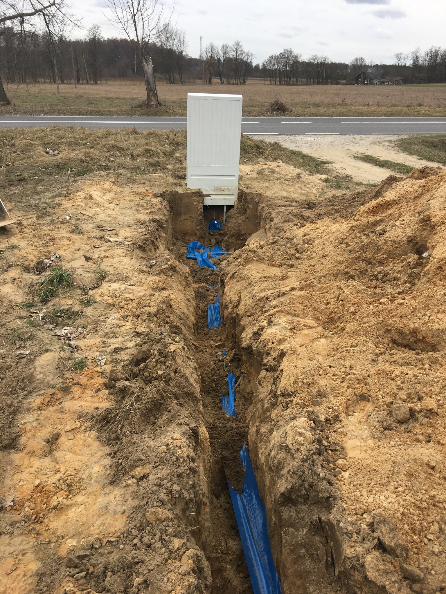 Wąski wykop w ziemi z niebieską folią zabezpieczającą, obok drogi i białej szafki elektrycznej na tle pola i drzew.