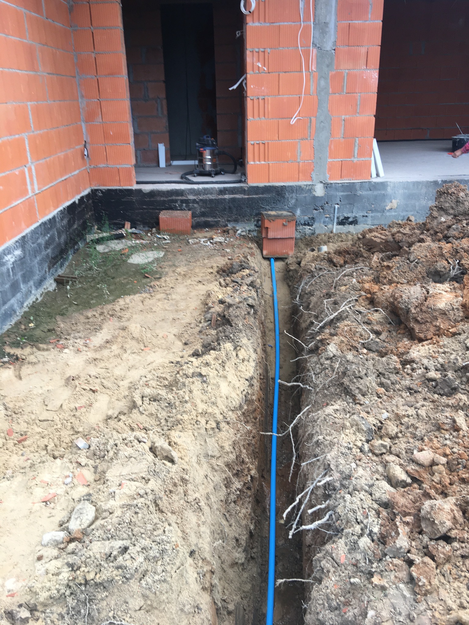 Wąski wykop z niebieską rurą prowadzącą do ceglanej studzienki przy fundamencie budynku z pustaków ceramicznych.