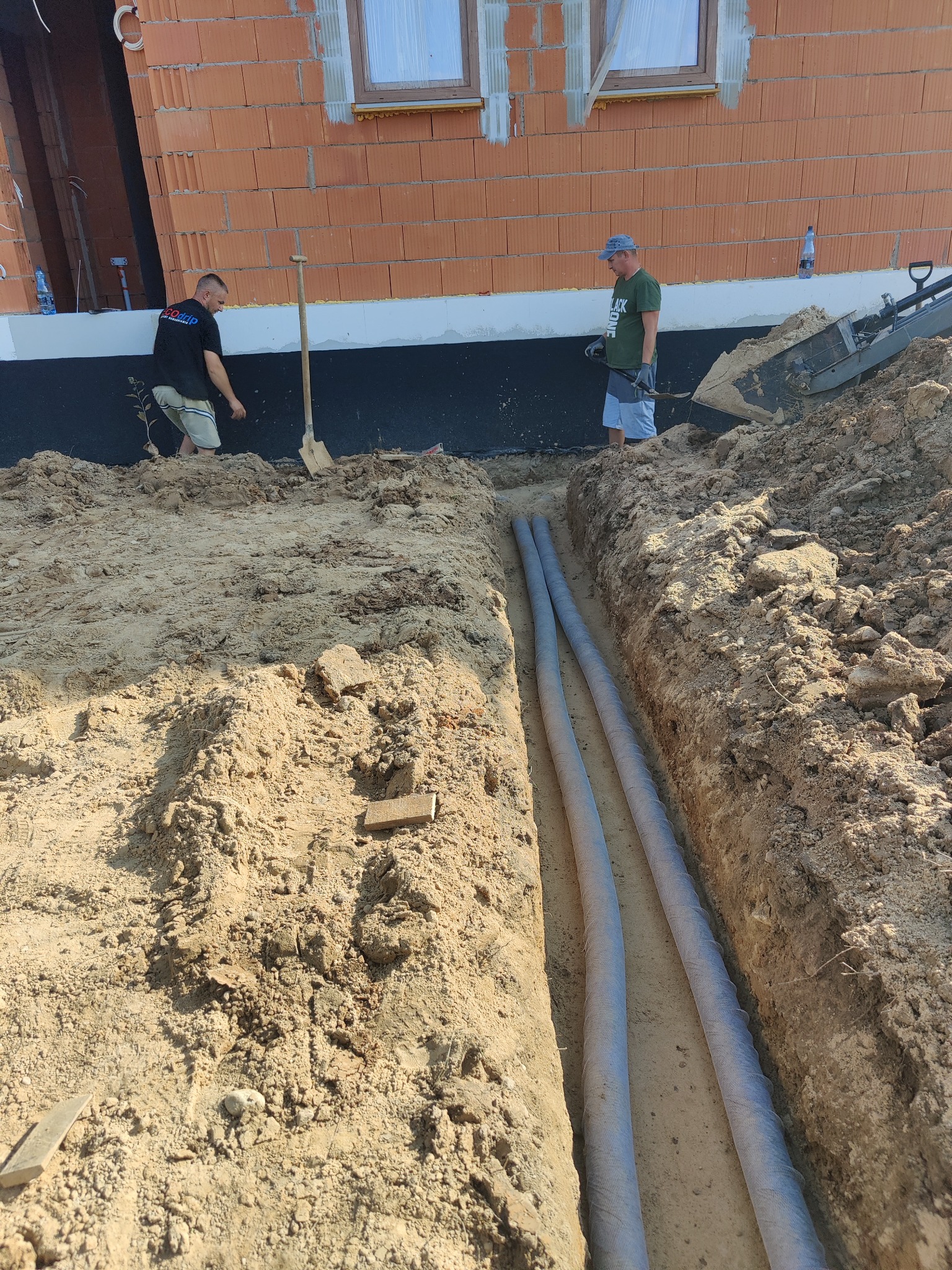 Dwaj mężczyźni pracują przy wykopie pod budynkiem z cegły, w którym ułożone są dwie rury w osłonie, widoczna łopata i sprzęt budowlany.