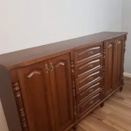 Klasyczna, długa komoda z ciemnego drewna z frezowanymi frontami szuflad i szafek, ozdobnymi uchwytami i rzeźbionymi detalami, ustawiona na drewnianej podłodze przy jasnej ścianie.