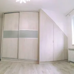 Biała szafa narożna z przesuwnymi drzwiami i szarym pasem, dopasowana do skosu dachu, na podłodze panele w odcieniu jasnego drewna.