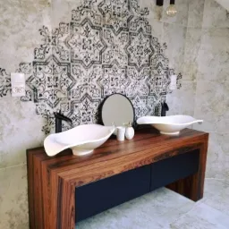 Umywalki nablatowe w kształcie łódek na drewnianej szafce z czarnymi szufladami, wzorzysta ściana w tle, wiszące żarówki na kablach.