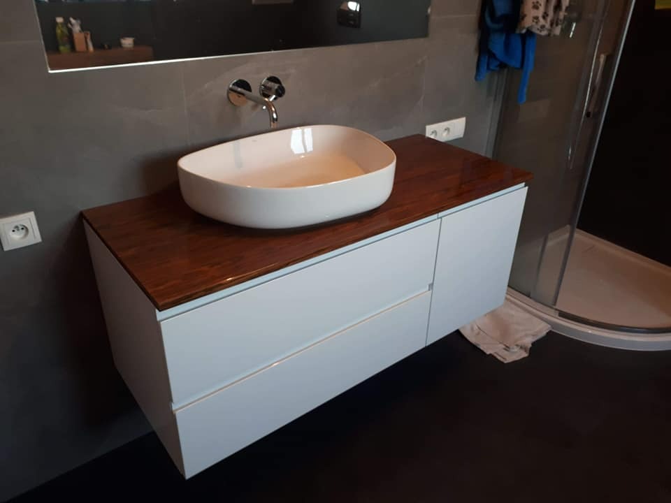 Nowoczesna łazienka z białą, owalną umywalką nablatową na szafce z białymi frontami i blatem z ciemnego drewna, z widoczną baterią ścienną i szarą ścianą w tle.