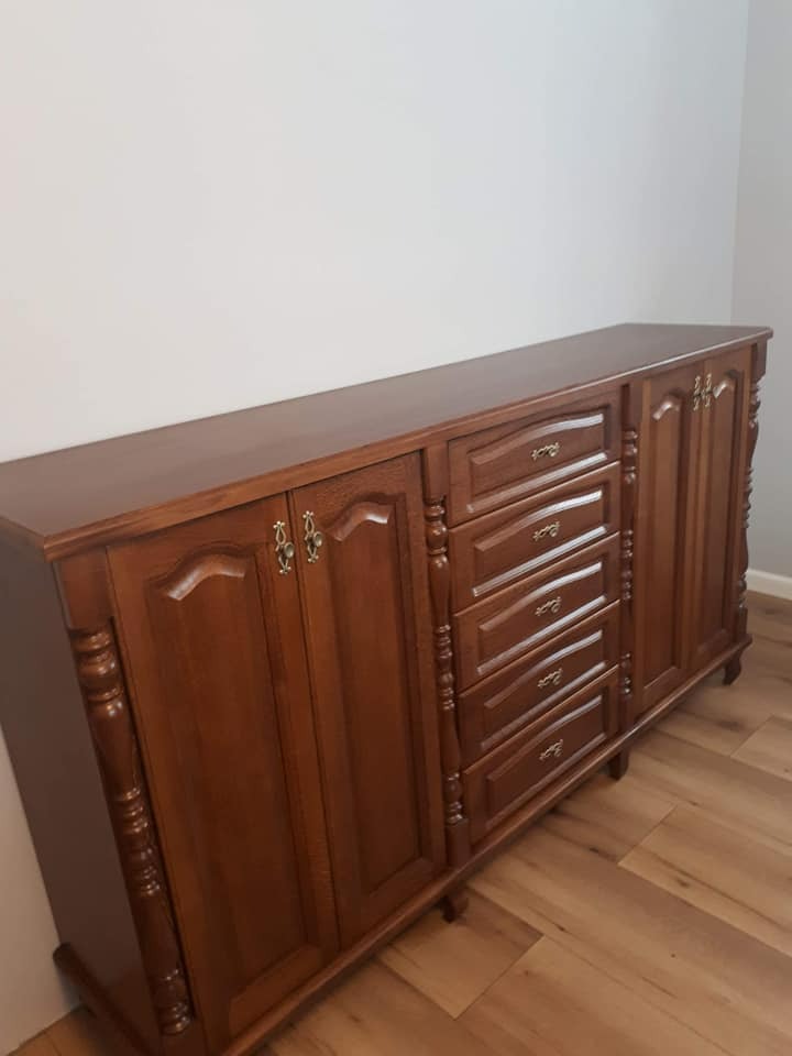 Klasyczna, długa komoda z ciemnego drewna z frezowanymi frontami szuflad i szafek, ozdobnymi uchwytami i rzeźbionymi detalami, ustawiona na drewnianej podłodze przy jasnej ścianie.