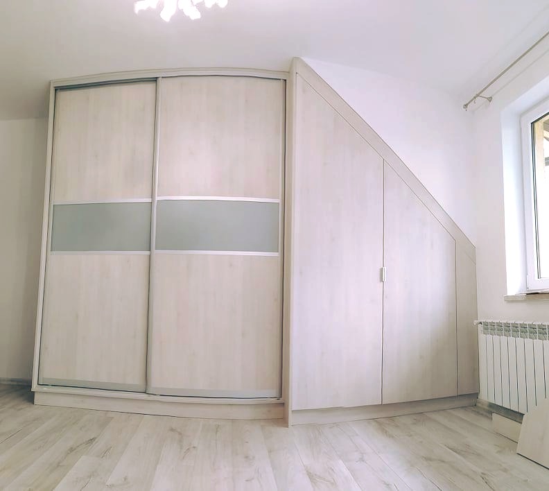Biała szafa narożna z przesuwnymi drzwiami i szarym pasem, dopasowana do skosu dachu, na podłodze panele w odcieniu jasnego drewna.
