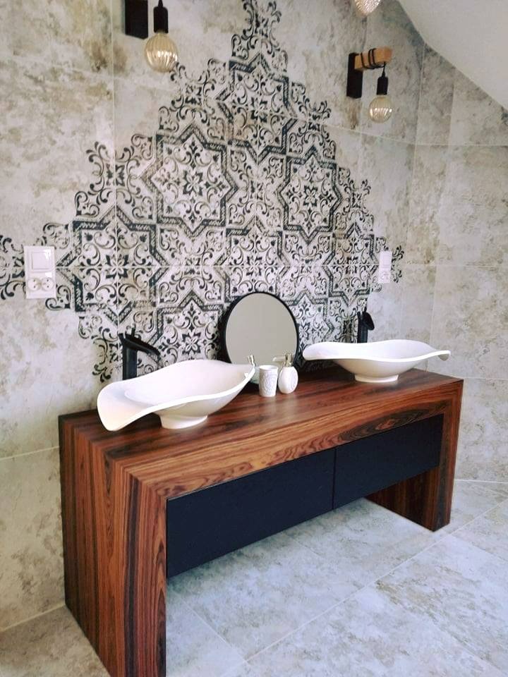 Umywalki nablatowe w kształcie łódek na drewnianej szafce z czarnymi szufladami, wzorzysta ściana w tle, wiszące żarówki na kablach.