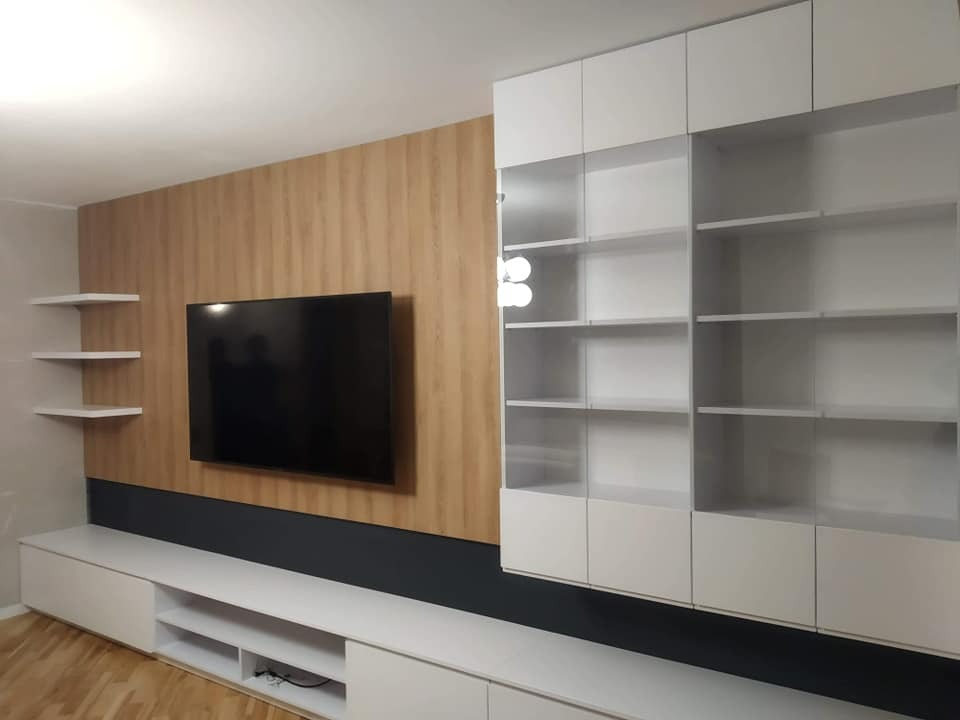 Biała zabudowa ściany z szafkami i półkami, z drewnopodobnym panelem ściennym i wiszącym telewizorem, minimalistyczny design.