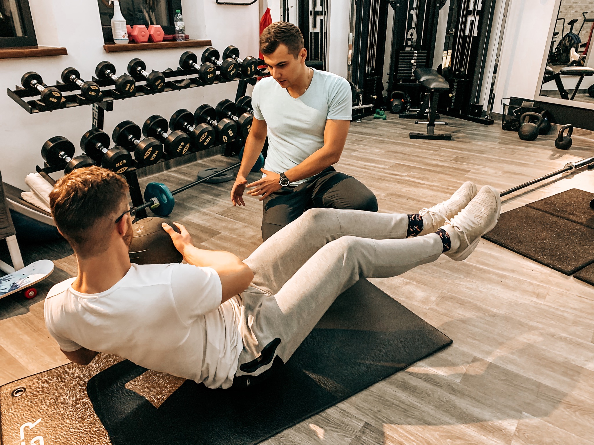 Mężczyzna wykonuje skłony z piłką lekarską na macie pod okiem instruktora w siłowni, w tle widoczny stojak z hantlami i sprzęt do ćwiczeń.