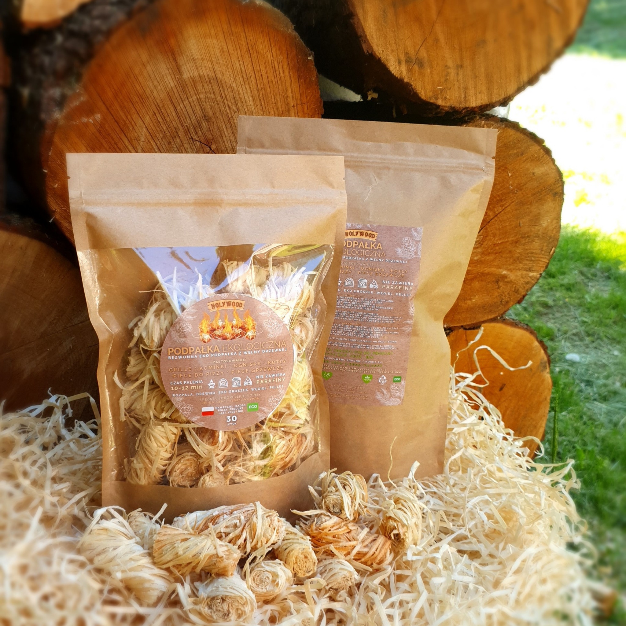 Dwa opakowania ekologicznej podpałki z wełny drzewnej 'Holywood' na tle stosu drewna kominkowego i dekoracyjnych wiórów.
