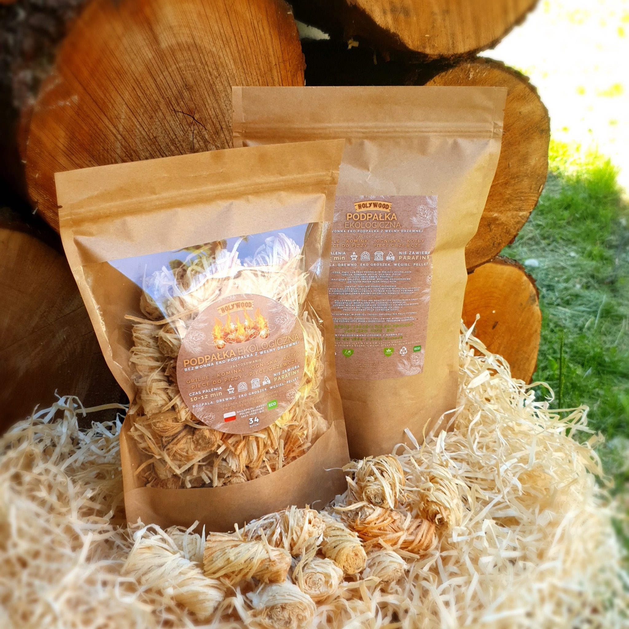 Dwa opakowania ekologicznej podpałki z wełny drzewnej 'Holywood' na tle ułożonych pniaków i wypełniacza z wiórów papierowych, z widocznymi informacjami o produkcie i polską flagą.