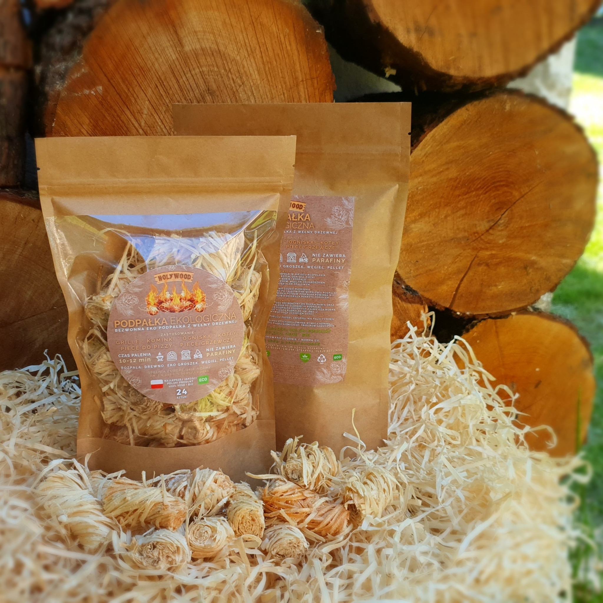 Dwa opakowania ekologicznej podpałki z wełny drzewnej, jedno przezroczyste, drugie w papierowym worku, ułożone na podpałce luzem, na tle stosu pociętych pni drzew