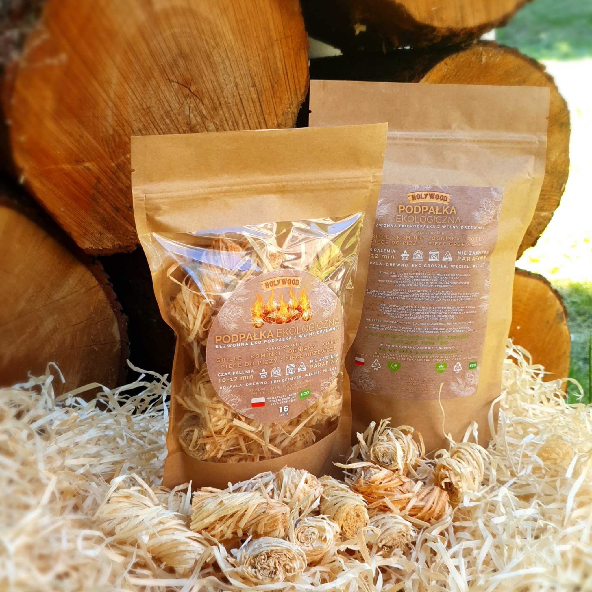 Dwa opakowania ekologicznej podpałki z wełny drzewnej, marki Holywood, ułożone na słomie na tle stosu drewna kominkowego.