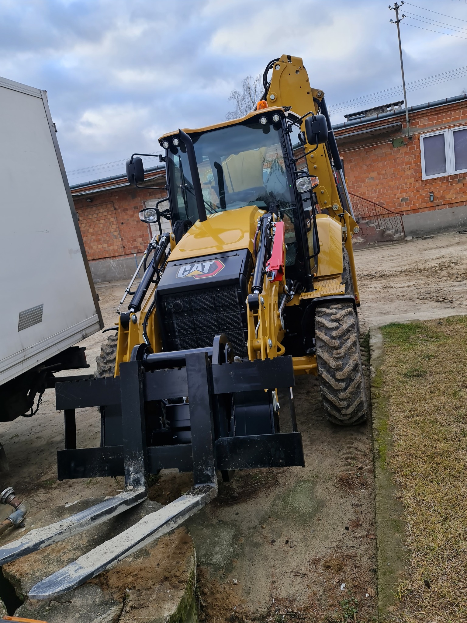 Żółta koparko-ładowarka CAT z widłami z przodu, stojąca na ziemi obok białej przyczepy, na tle ceglanego budynku z oknami.