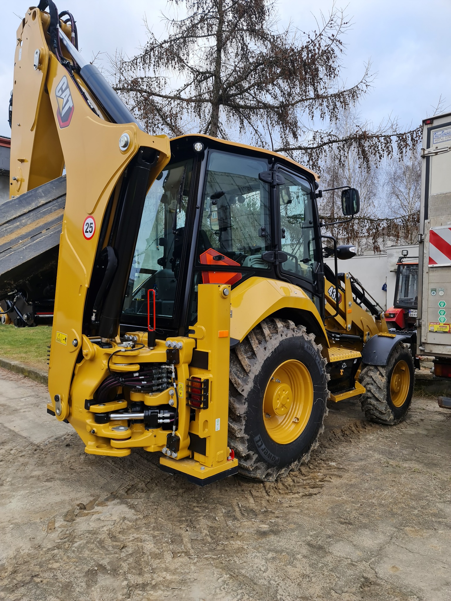 Żółta koparko-ładowarka CAT 434 z widocznymi śladami błota na oponach i ziemi, zaparkowana obok białej przyczepy, na tle drzewa z brązowymi liśćmi.