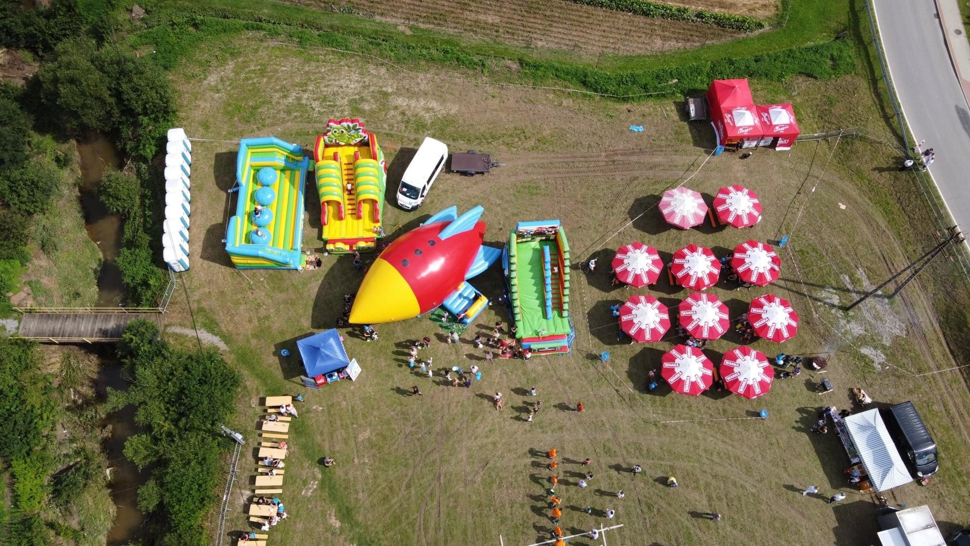Widok z lotu ptaka na teren pikniku z różnorodnymi atrakcjami, w tym kolorowe dmuchane zamki, zjeżdżalnie w kształcie rakiety, strefa relaksu z parasolami i stolikami piknikowymi, oraz potok...