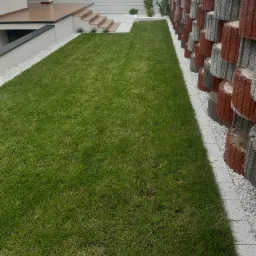 Nowoczesny ogród z równo przystrzyżonym trawnikiem, białymi kamykami oddzielającymi trawnik od ściany z betonowych donic w kolorach szarości i czerwieni, oraz białym ogrodzeniem w tle.