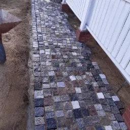 Ścieżka z kostki granitowej w trakcie układania, widoczny wózek budowlany i fragment budynku z białymi panelami.