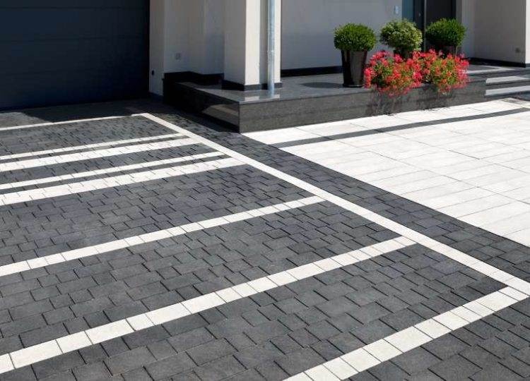 Nowoczesny podjazd z szarej kostki brukowej z białymi pasami, prowadzący do domu z ciemną bramą garażową i ozdobnymi roślinami w donicach.