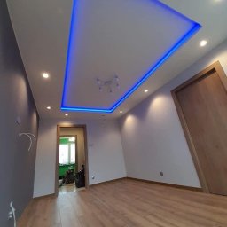 Gambud - Nowoczesne wykończenie wnętrza pokoju z drewnianą podłogą, białymi ścianami, szarą ścianą akcentującą i podwieszanym sufitem z niebieskim oświetleniem LED.