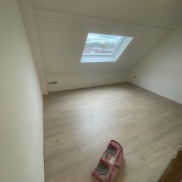 Gambud - Poddasze po remoncie z nową podłogą imitującą drewno, widoczne okno dachowe z widokiem na miasto i narzędzie do układania płytek podłogowych.