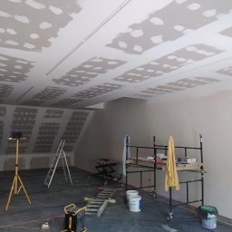 Gambud - Sufit w trakcie szpachlowania z widocznymi łączeniami płyt gipsowo-kartonowych, oświetlenie robocze na statywie, drabina i rusztowanie w pomieszczeniu.