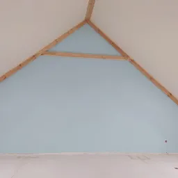 Pomalowana na błękitno ściana szczytowa poddasza z widoczną drewnianą konstrukcją więźby dachowej i wystającymi przewodami elektrycznymi z sufitu.