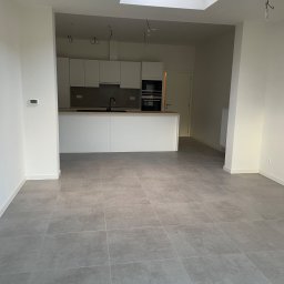 Gambud - Nowoczesne wnętrze z szarymi płytkami podłogowymi, białą kuchnią w tle i świetlikiem sufitowym, widoczne niedokończone instalacje elektryczne.