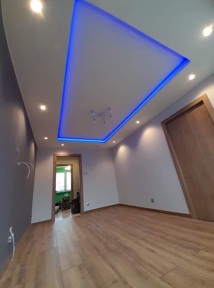 Nowoczesne wykończenie wnętrza pokoju z drewnianą podłogą, białymi ścianami, szarą ścianą akcentującą i podwieszanym sufitem z niebieskim oświetleniem LED.