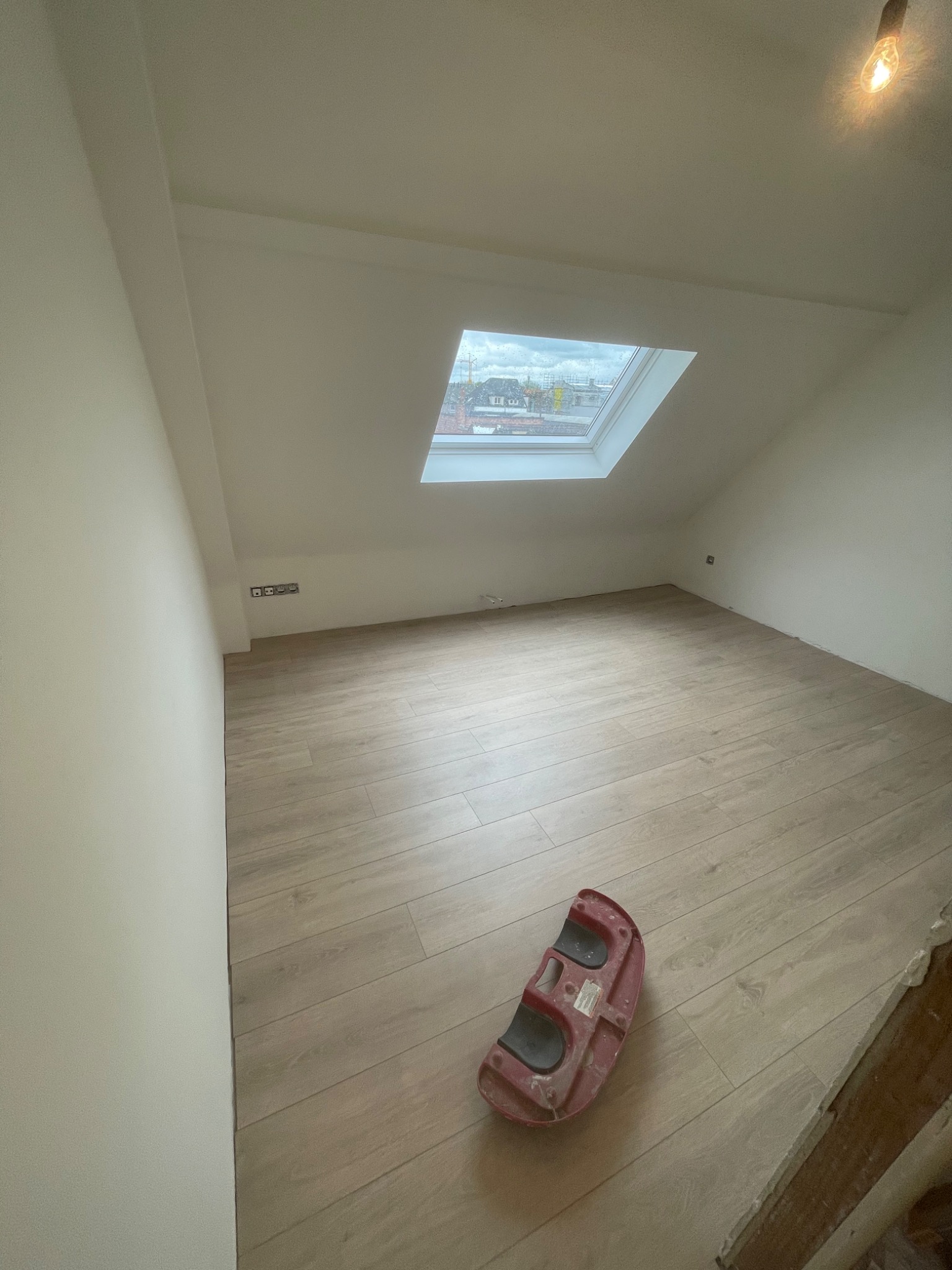 Poddasze po remoncie z nową podłogą imitującą drewno, widoczne okno dachowe z widokiem na miasto i narzędzie do układania płytek podłogowych.