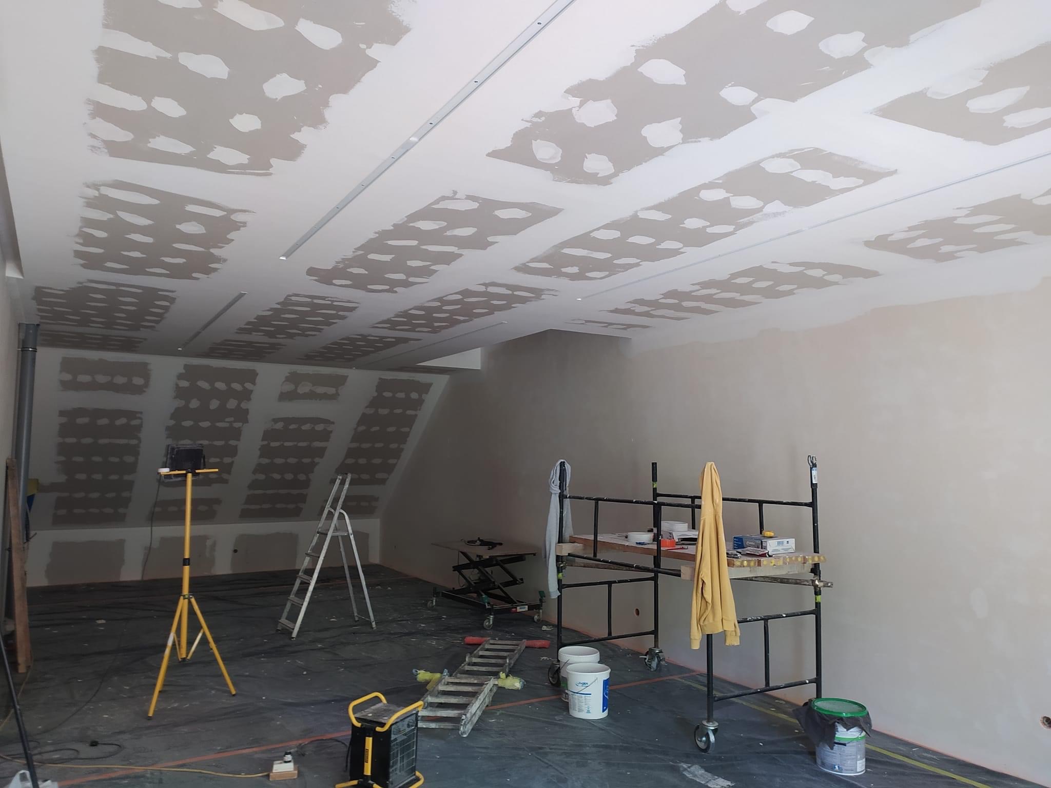 Sufit w trakcie szpachlowania z widocznymi łączeniami płyt gipsowo-kartonowych, oświetlenie robocze na statywie, drabina i rusztowanie w pomieszczeniu.